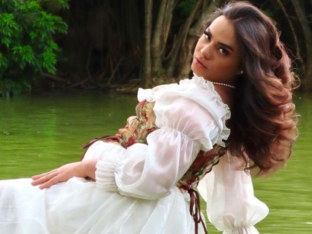LiliithBuckland with wavy brown hair, wearing a white, frilled, Victorian-style dress with floral corset, pearl necklace, and looking over her shoulder, standing by green water.