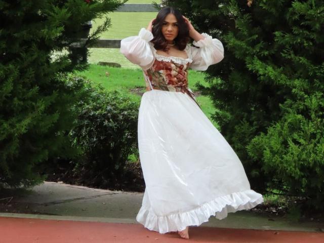LiliithBuckland with wavy brown hair, wearing a white, puffy-sleeve dress with a floral corset, standing between green trees, hands in hair, white frilled hem, red pavement.