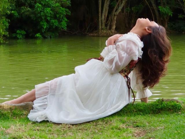LiliithBuckland is leaning back with her head tilted up, long brown hair flowing, wearing a white dress with a brown corset, her arms crossed over her tits, by a green pond.