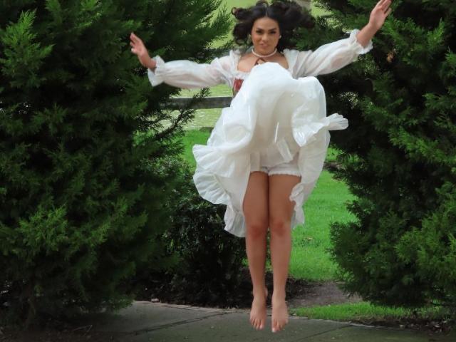 LiliithBuckland is mid-air, legs together, arms spread, wearing a white dress, flared out, between two green evergreen trees, barefoot, looking forward.