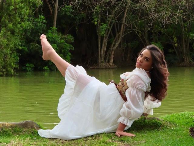 LiliithBuckland is lying on grass by a green pond, wearing a white, ruffled dress, with one leg raised, showing her bare foot, and a brown belt.