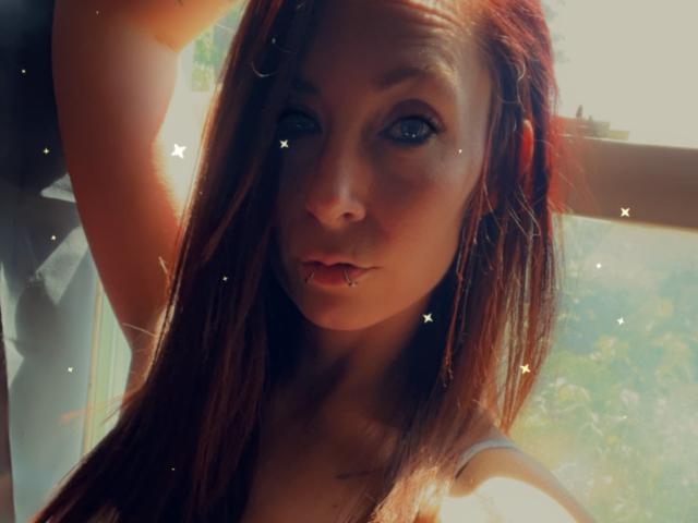 BeeHave69 has long brown hair, blue eyes, and a pierced lower lip. Sunlight highlights her face, with a slight shadow on her right cheek. Sparkles are scattered across the image.