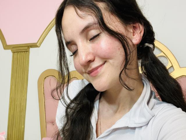 ChloeDulce, con le trecce, una camicia bianca e un sorriso carino, che mostra il suo piccolo naso e le lentiggini. Il suo seno non è visibile. Sta guardando in basso con gli occhi chiusi..