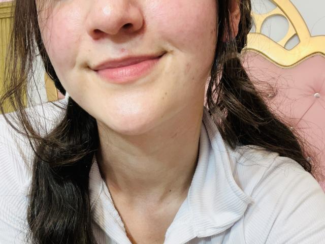 Il viso di ChloeDulce, con un piccolo sorriso, guance rosate, pelle chiara, capelli castani ondulati, e indossa una maglietta bianca a coste, con una piccola lentiggine sul mento..