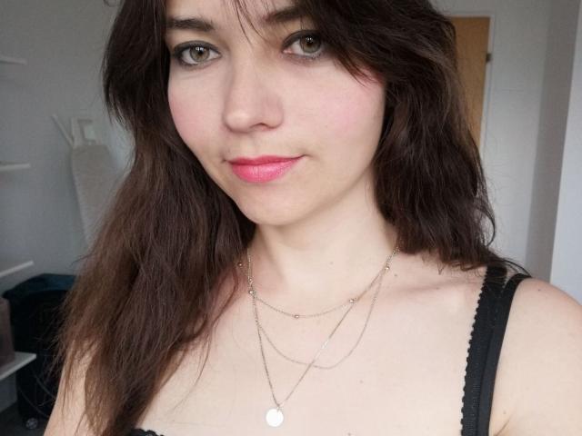 CyndeeWinter with wavy brown hair, light skin, green eyes, pink lips, black tank top, and two silver necklaces showing a small round pendant. Her tits are visible.