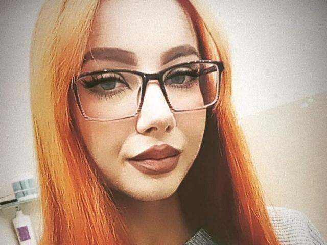 ValStephie with red-orange hair, wearing black-rimmed glasses, has full lips and a slight smile. Her large tits are partially visible under a grey shirt.