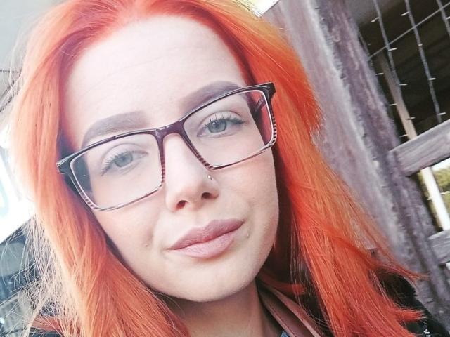 ValStephie has bright orange hair, wears dark glasses, and has a nose piercing with a small stud. Her lips are puckered in a slight pout.