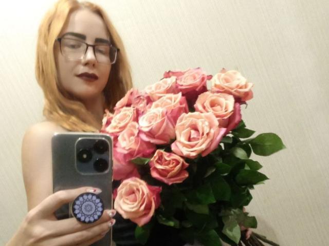 ValStephie with blonde hair, glasses, and dark lipstick, holding a bouquet of pink roses in front of a mirror, taking a selfie with a grey phone.