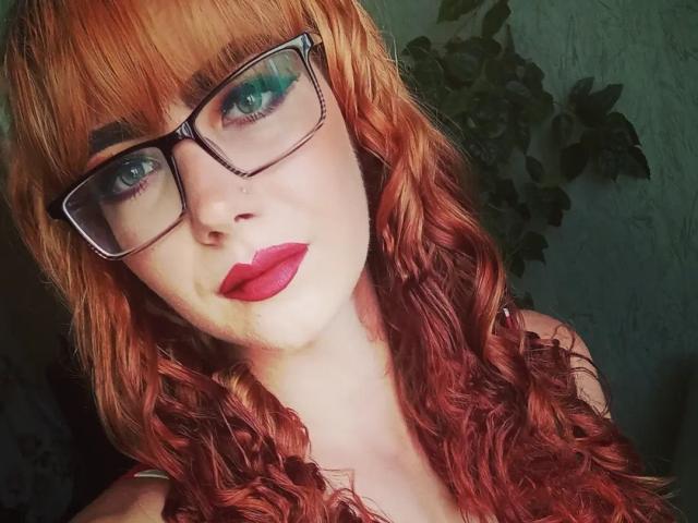 ValStephie with red curly hair, green eyes, and black glasses, wearing bold red lipstick, looking directly at the camera. Her tits are partially visible.