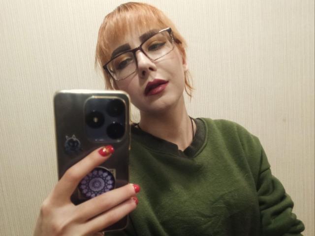 ValStephie with red hair, glasses, and red lipstick, wearing a green sweater, taking a selfie with a phone, red nails, slight smirk, textured wall in background.