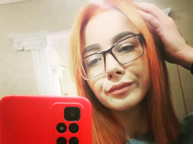 ValStephie with red hair, glasses, and a smile, holding a red phone, hand in hair, showing her face and phone in a selfie. Her tits are not visible.