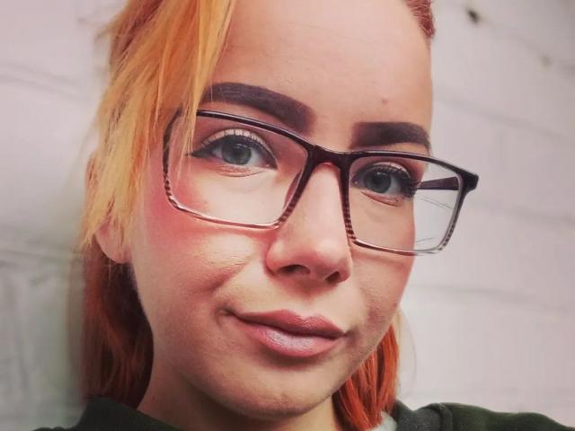 ValStephie with orange hair, thick eyebrows, and dark glasses, showing a subtle smile, with a slight blush on her cheeks, wearing a green shirt.