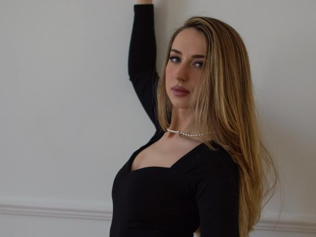 JulietteCute with long blonde hair, black top, and pearl necklace, arms raised, looking at camera with a subtle smile, showing her tits and ass in the photo.
