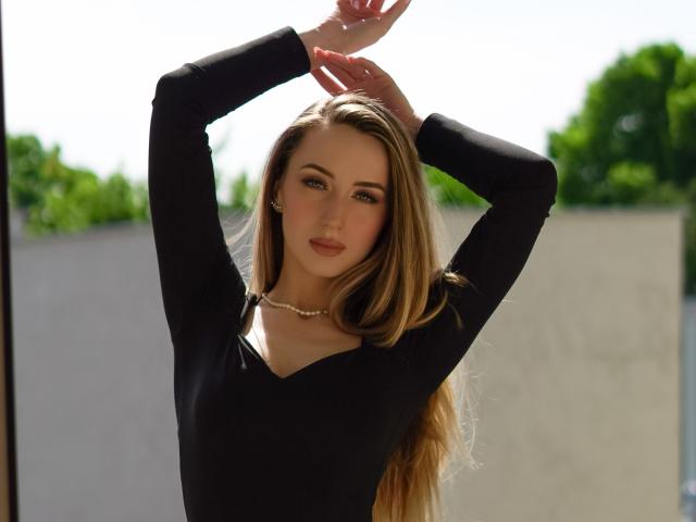 JulietteCute has long blonde hair, wearing a black long-sleeve top, arms raised, pearl necklace, and a subtle smile, with blue eyes and pink lipstick, looking sexy.