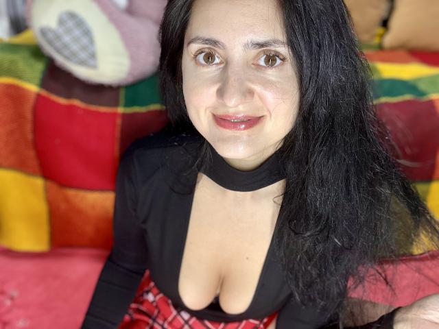 JennaEasy with long black hair, brown eyes, and a slight smile, wearing a deep black top revealing her big boobs and a red plaid skirt, sitting on a colorful blanket.