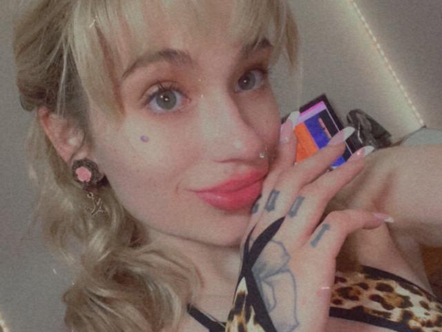 MarryJaneXO with blonde hair, pink lipstick, and tattoos, holding a phone, wearing leopard print, pink earbuds, and a nose ring, smiling with pink lips.