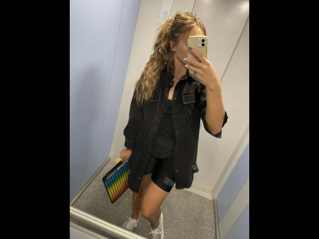 GanzNettePaola-hot with wavy blonde hair, black oversized shirt, black shorts, white sneakers, holding colorful checkered bag, taking mirror selfie in a lit, white-walled room.