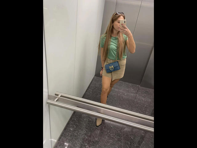 GanzNettePaola-hot with long blonde hair, green top, beige mini-skirt, blue crossbody bag, and tan boots, taking a mirror selfie in a elevator, showing her ass.