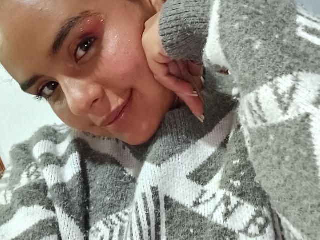 ArielleWilde with a cute smile, her head resting on her hand, wearing a gray and white patterned sweater, brown eyes with subtle eye makeup, and shiny skin.