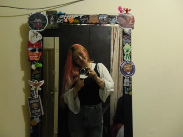 IsabellaClown with pink hair, white sleeves, black top, gray pants, holding camera in mirror, surrounded by stickers, smiling. Her ass is visible in the mirror reflection.