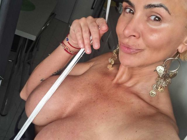 BriliantOne with tan skin, brown eyes, and short dark hair, holding a pole, topless, showing large tits, wearing gold earrings, and red bracelets, smiling slightly.