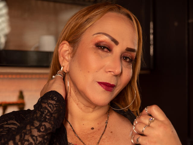 DianeOasis, a blonde woman with red lipstick and black lace top, holds her ear, showing off rings and hoop earrings, with a slight smile, looking sexy.