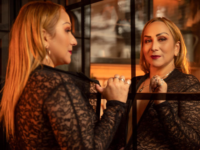 DianeOasis with blonde hair, wearing a black lace top, looking in a mirror, holding a black hairbrush, showing off her red lipstick and rings, in a warm, orange-lit room.