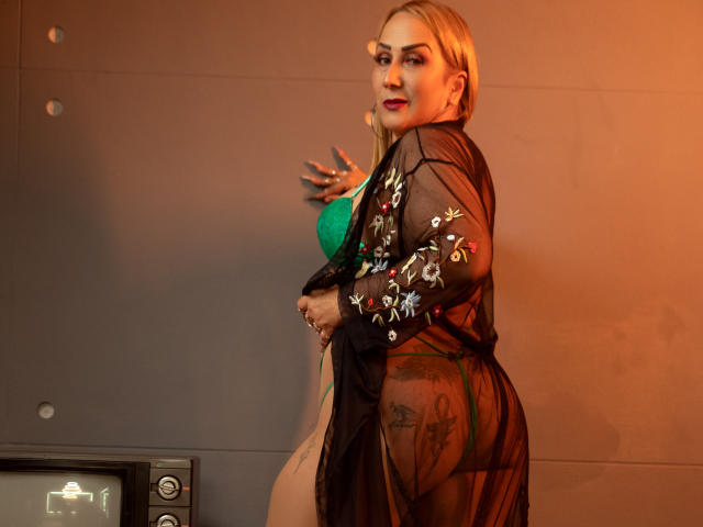 DianeOasis, avec ses cheveux blonds, son soutien-gorge vert et sa robe noire transparente, exposant ses seins et ses fesses, avec un tatouage visible sur sa joue droite, se tient devant une lumière orange chaude..