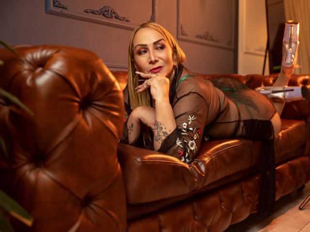 DianeOasis, blonde, lying on a brown leather couch, wearing a sheer black robe, showing tattoos on her arm, resting her chin on her hand, looking at the camera.