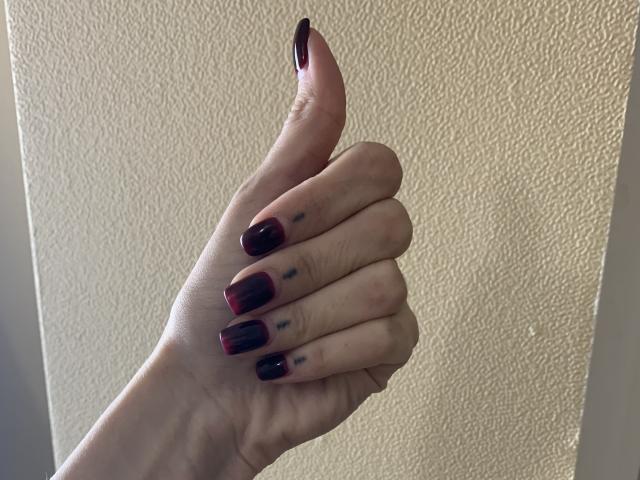 MoonLiza's hand with dark red, glossy nails, giving a thumbs-up. Small black tattoos on fingers. Light skin, textured beige wall in background. MoonLiza's thumb is most prominent.