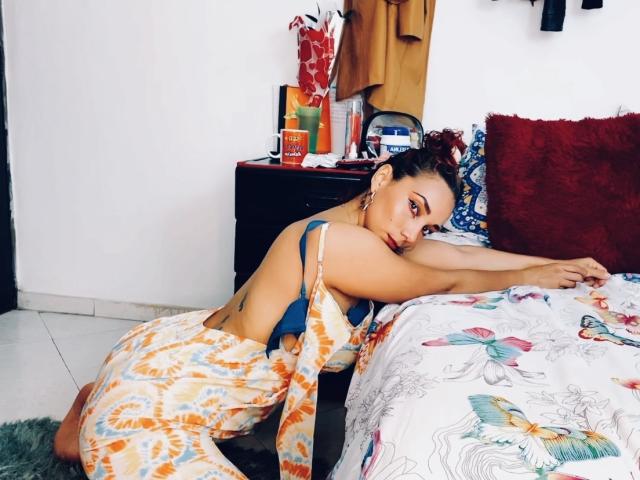 AmberDemons with tan skin and dark hair in a bun, wearing a yellow-orange patterned dress, leaning on a bed with colorful butterfly-patterned sheets, exposing her ass.