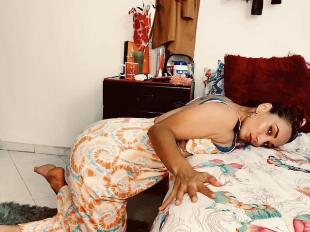 AmberDemons is on a bed, wearing a tie-dye robe, looking back with red eyes, her hand on the bed. Her ass is slightly raised, showing her butt.