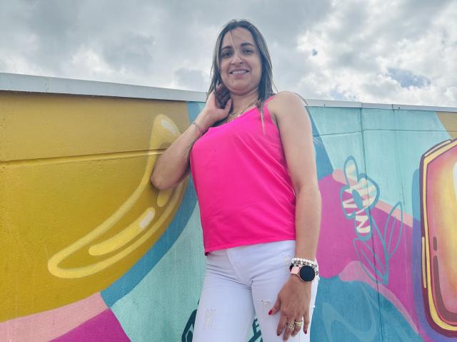 AliceDuboiss with long brown hair, pink tank top, white pants, and gold bracelet, standing against colorful graffiti wall, smiling, right hand touching her hair.