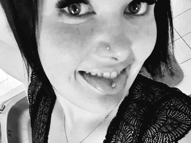 Black-and-white selfie of Chafouine with a nose ring, tongue piercing, and a lip piercing, wearing a lace top, looking up with a slightly open mouth, showing teeth.