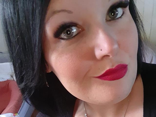 Chafouine has dark hair, green eyes with heavy black eyeliner, red lipstick, a small nose piercing, and a slight smile, wearing a black top, in a selfie.
