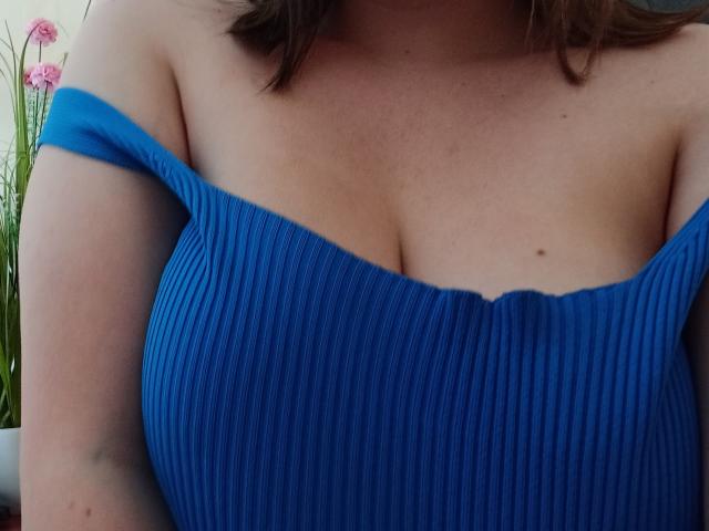 DiLaria's blue ribbed off-shoulder top shows her big, round tits with a cute mole above. Her brown hair frames her chest, and pink flowers are in the background.