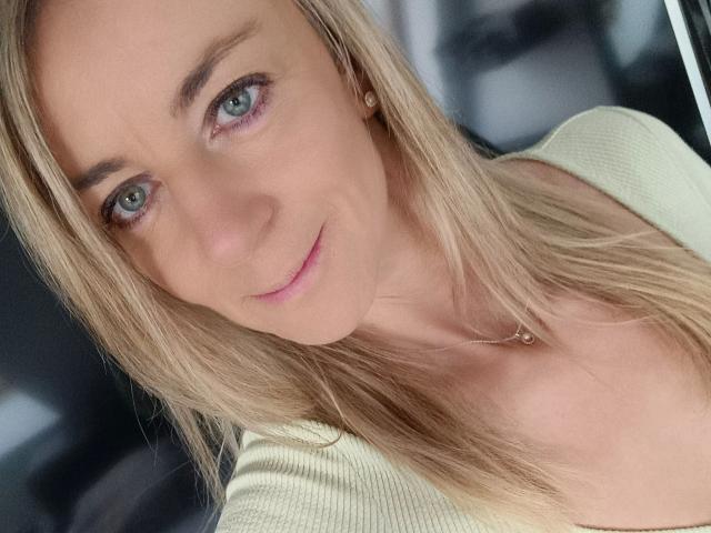 LissLonglegs-hot has blonde hair, blue eyes, and a subtle smile. She's wearing a green top, and her small, perky tits are partially visible. Her skin is fair.