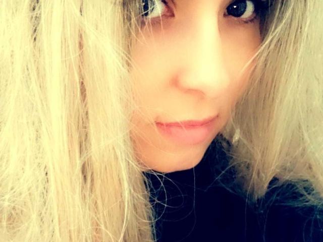 LucyELY's face with blonde, slightly messy hair covering one eye. She has pale skin, small pink lips, and dark, curious eyes. She's wearing a black turtleneck.
