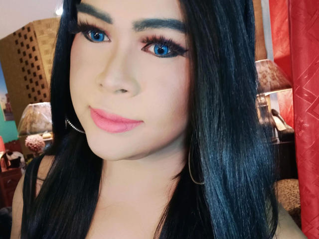 transsweetdenise-hot with blue eyes, black hair, pink lipstick, long eyelashes, hoop earrings, smiling, fair skin, makeup, and a cute face. Boobs partially visible in black top.