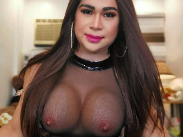Transsweetdenise-hot with long brown hair, pink lipstick, and hoop earrings, wearing a black sheer top, exposing her big, shiny, bare tits with small, erect nipples.