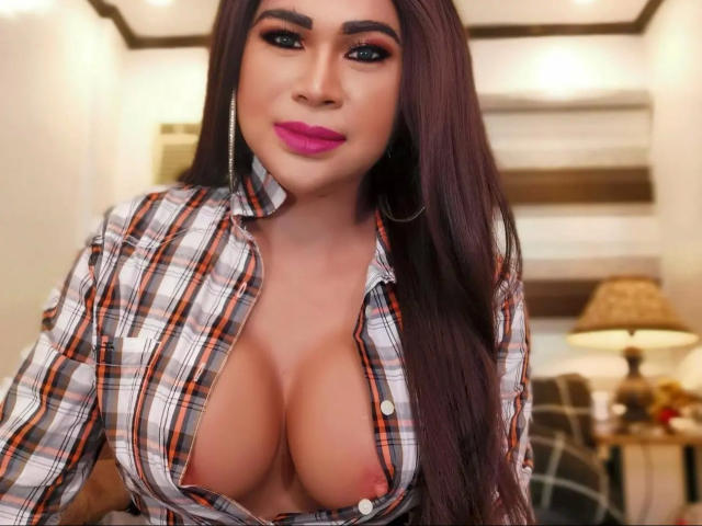 transsweetdenise-hot with long brown hair, pink lipstick, and hoop earrings, wearing an open plaid shirt, exposing large, round, tan tits, standing in a warmly lit room.