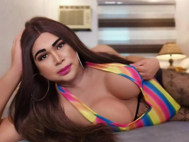 Transsweetdenise-hot with long brown hair, pink lipstick, hoop earrings, and rainbow-striped top, showing big tits in a low-cut top, lying on a bed, looking at the camera.