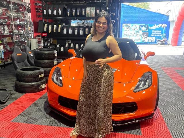 BrianaPreston with big tits, wearing a black crop top and a long leopard print skirt, standing in front of a shiny orange sports car in a tire shop.