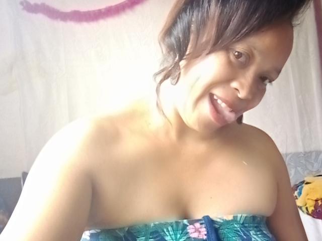 Rahel69, con la pelle scura e i capelli neri, sorride, mostrando le spalle scoperte e piccoli seni, indossando un top senza spalline con un motivo verde e blu. Luce sullo sfondo..