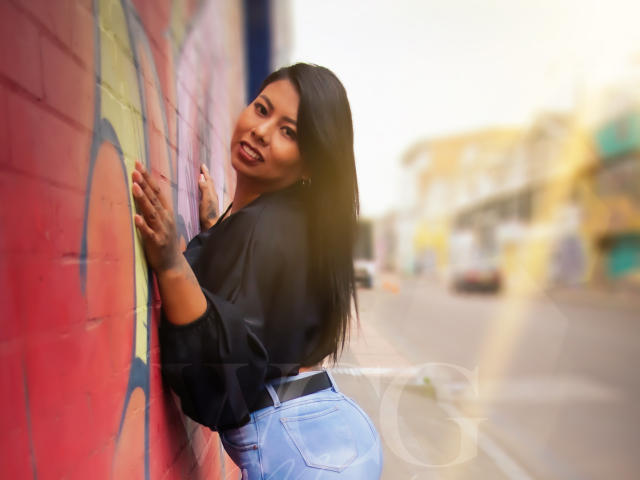 LizHerrera with long black hair, smiling, leaning against a red wall, wearing a black top and blue jeans, showcasing her round ass, standing on a sunny street.