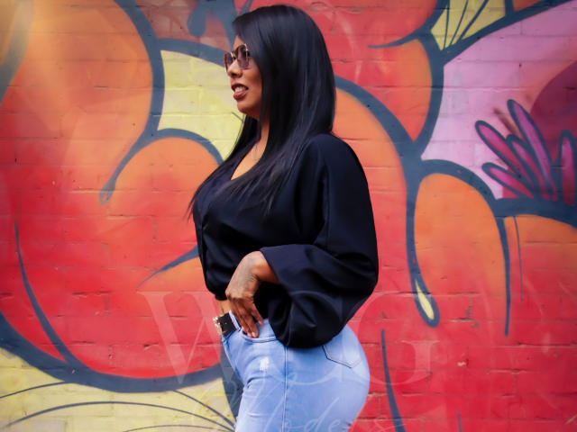LizHerrera with long black hair, wearing a black blouse and blue jeans, standing sideways, showing her big ass, in a colorful graffiti wall background, wearing sunglasses.