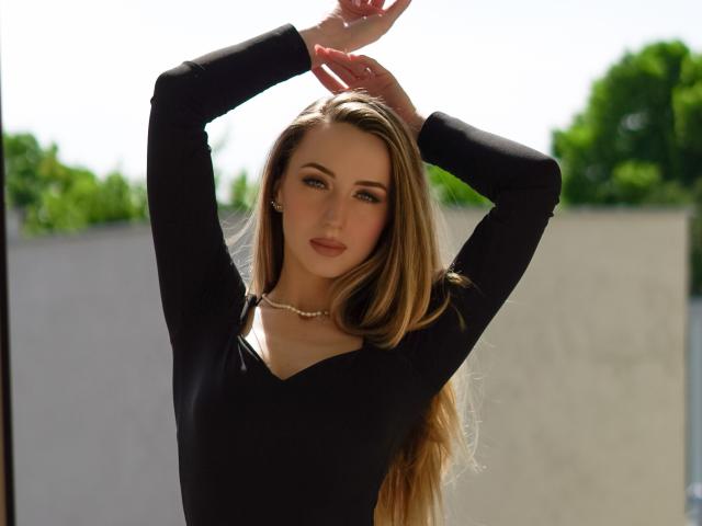 JulietteCute has long blonde hair, wearing a black long-sleeve top, arms raised, hands touching her head, with a pearl necklace, and a sexy, confident expression.