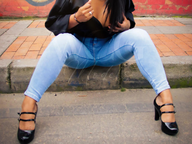 LizHerrera is sitting with legs spread, wearing light blue jeans, black high heels, and a black top, exposing her tits. She's holding her top down with one hand.