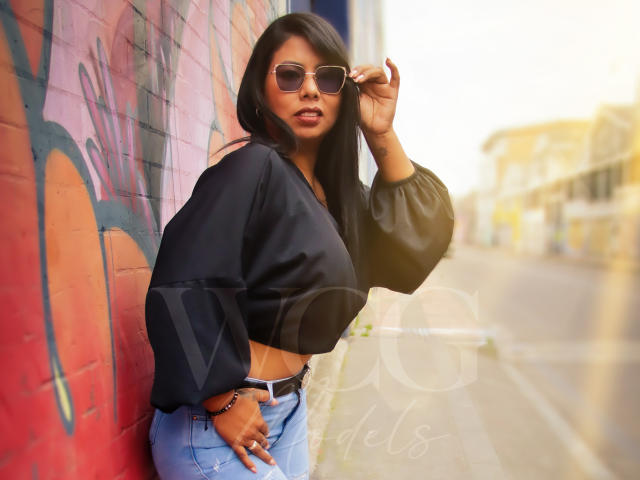 LizHerrera with big tits, wearing a black crop top and blue jeans, standing against a graffiti wall, adjusting her sunglasses, with long black hair and a sexy look.