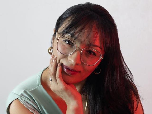 LexiLuna with glasses, holding her cheek, green top, long black hair, red light, gold hoop earrings, subtle smile, light skin, small nose, small pink lips, slightly blushing.